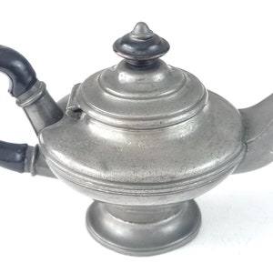 Op de afbeelding: Een klein tinnen theepotje met een zwart handvat en een gewelfd deksel. De theepot heeft een rond lichaam en een uitlopende tuit.
