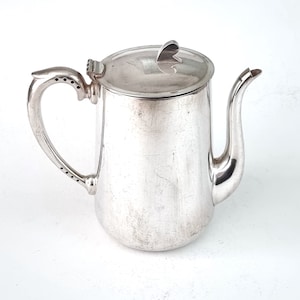 May include: A silver teapot with a heart-shaped lid handle. The teapot has a smooth, polished surface and a slightly curved spout.
