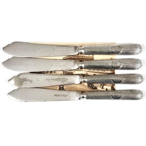 Puede incluir: Un juego de cinco cuchillos de mantequilla antiguos. Cada cuchillo tiene una hoja plateada ligeramente curvada y un mango acanalado. Los cuchillos están dispuestos sobre un papel envejecido con una imagen descolorida.