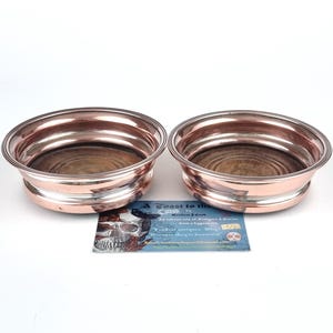 May include: Two copper-toned coasters with silver rims and wooden centres. The coasters are circular and low-profile. A business card with a skull and text is visible between the coasters.