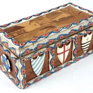 Puede incluir: Una caja de madera con un diseño decorativo. La caja está pintada con acentos azules, rojos y blancos. La parte superior de la caja presenta una escena de un pueblo con casas y una iglesia. Los lados de la caja están decorados con escudos.