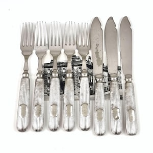 Puede incluir: Un juego de cubiertos antiguos de plata, que incluye tenedores y cuchillos de mantequilla. Los utensilios plateados tienen mangos acanalados con un emblema en forma de escudo. Los tenedores tienen cuatro púas. Los cuchillos tienen una punta redondeada. El juego está sobre un fondo blanco.