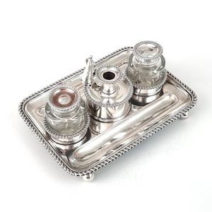 May include: A silver-plated desk set with a rectangular tray and three glass inkwells. The set includes a central ink pot, two inkwells, and a pen rest. The tray has a rope-like border and rests on small ball feet.