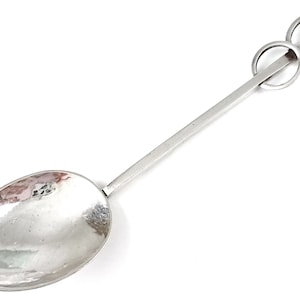 Puede incluir: Una cuchara de plata con un diseño de mango único que presenta dos anillos entrelazados. La cuchara tiene un cuenco liso de forma ovalada.