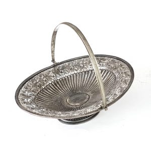 Puede incluir: Una cesta de servir ovalada de plata con un diseño floral decorativo y un asa. La cesta tiene un centro elevado y un borde festoneado.