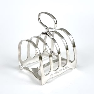 May include: A silver-plated toast rack with a handle and four compartments. The rack has a curved design and the words "CRANAGE CAFE" are engraved on the base. The rack is made of metal and sits on a white surface.