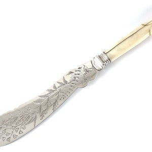 Puede incluir: Un cuchillo de mantequilla de plata con mango de hueso. La hoja está decorada con un diseño floral grabado.