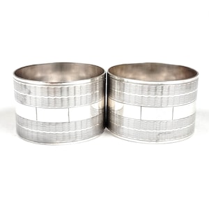 May include: Two silver napkin rings. Each ring features a textured, wavy pattern at the top and bottom, with a smooth, rectangular band in the middle. The rings are made of a shiny, reflective metal.