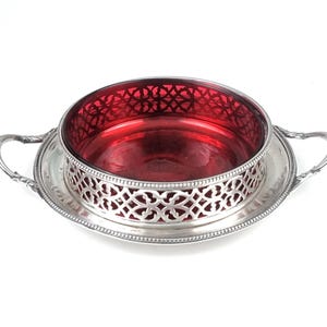 May include: An antique serving dish with a ruby red glass bowl and a silver-plated filigree design. The dish has a round base with two handles. The bowl is set in a decorative silver frame.