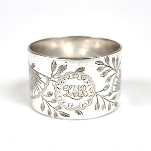 May include: A silver napkin ring with engraved floral and leaf patterns. The ring has an elaborate central cartouche with the initials "LMB". The ring is cylindrical and made of a shiny, reflective metal.