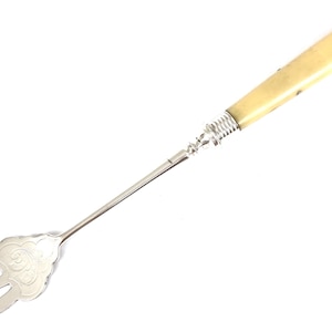 May include: A silver serving fork with a long, slender handle made of ivory. The fork has a decorative, ornate design and three prongs.