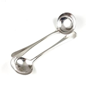 May include: Two shiny, silver ladles with rounded bowls and long, slender handles are displayed against a white background. The ladles are angled, with one resting atop the other. The reflective surfaces suggest a polished finish, suitable for serving soups or sauces.