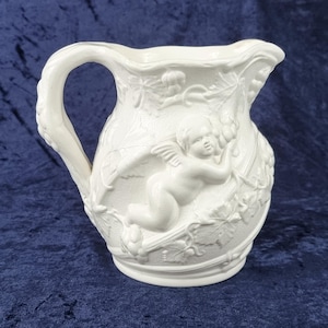 May include: White ceramic pitcher with a detailed relief design. The pitcher features a cherub, grape vines, and leaves. The handle is curved and the spout is gently flared. The pitcher is a decorative item.