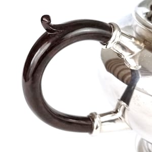 May include: Close-up of a silver teapot with a black, curved handle. The handle is attached to the teapot with a silver band.