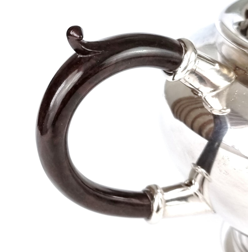 May include: Close-up of a silver teapot with a black, curved handle. The handle is attached to the teapot with a silver band.