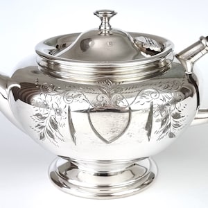 May include: A silver teapot with an ornate engraved design and a black wooden handle.