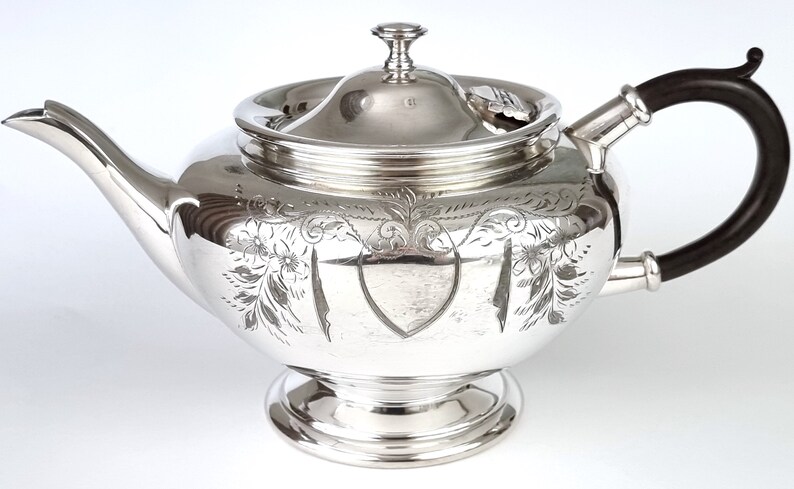 May include: A silver teapot with an ornate engraved design and a black wooden handle.