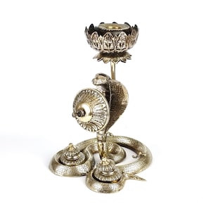 May include: A silver-colored candle holder with a cobra design. The candle holder features a cobra head and body, with a lotus flower-shaped holder at the top. The base has two decorative elements.