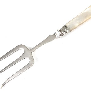 May include: A silver serving fork with a mother-of-pearl handle. The fork has three tines and is designed for serving food.