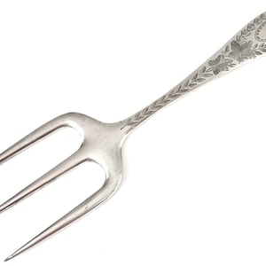 May include: A silver three-pronged fork with an ornate handle. The handle is decorated with a floral design and a blank oval.