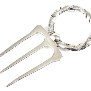 Könnte beinhalten: Eine silberfarbene Serviergabel mit drei Zinken und einem dekorativen Ringgriff. Der Griff hat ein florales Design. Die Gabel ist zum Servieren von Speisen bestimmt.
