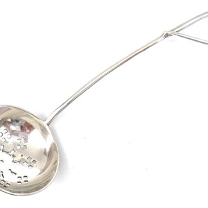 May include: Silver tea strainer with a round bowl and a long handle that ends in a loop. The bowl has a decorative pattern of small holes.