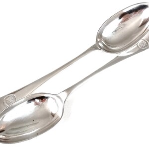 May include: Two silver spoons with long, slender handles and oval bowls are displayed against a white background. Each spoon has a small, circular emblem at the end of the handle. The spoons are angled, creating a sense of depth.