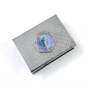 May include: A rectangular silver-toned metal case with a textured cross-hatch surface. Centered on the lid is a hexagonal enamel emblem featuring a light blue background, a green ship illustration, and the text G.M.V. ARAMOANA N.Z. RAILWAYS.