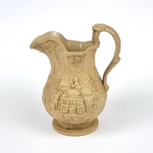 Puede incluir: Jarra de cerámica de color beige con un diseño en relieve detallado. La jarra tiene un pico, un asa curva y una base redonda. El relieve representa vides, uvas y figuras con un carro. Una pieza decorativa para la cocina o el comedor.