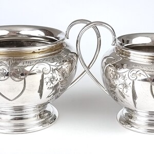 May include: A silver sugar bowl and creamer set with ornate floral designs. The pieces have a polished finish and a rounded shape.