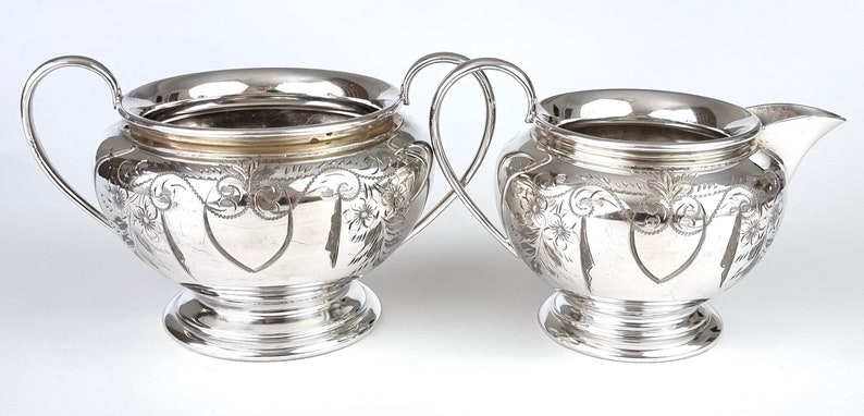 May include: A silver sugar bowl and creamer set with ornate floral designs. The pieces have a polished finish and a rounded shape.