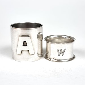 Könnte beinhalten: Zwei silberne Serviettenringe. Ein Ring ist zylindrisch mit dem Buchstaben "A" auf der Vorderseite und einem kleinen Griff. Der andere Ring ist kleiner, mit dem Buchstaben "W" auf der Vorderseite.
