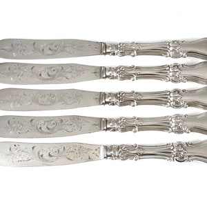 Può includere: Set di cinque coltelli da burro in argento con manici decorati e motivi floreali incisi.