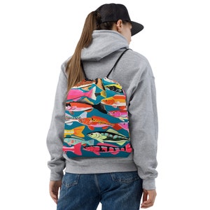 May include: A drawstring backpack featuring a vibrant pattern of colorful fish against a teal background. The fish vary in size and color, including pink, orange, yellow, and green. The backpack has black drawstrings and is worn over the shoulders.
