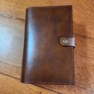 May include: A brown leather wallet with a flap closure and a single snap button. The wallet has a stitched edge and a smooth, textured surface.