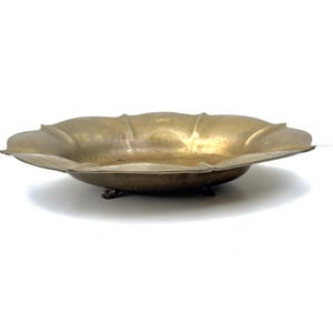 May include: A shallow, circular brass bowl with a scalloped edge featuring eight distinct lobes. The metallic surface has a warm, aged patina with visible variations in tone. The bowl is supported by three small, dark, integrated metal feet.