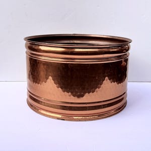May include: A copper-colored planter with a hammered texture. The cylindrical container has a rolled rim and two horizontal bands. The interior is dark, and the exterior reflects light, creating a warm, metallic appearance. A decorative piece for indoor or outdoor use.