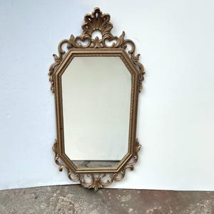 May include: Ornate gold-colored mirror with an octagonal frame. The mirror features intricate scrollwork and a decorative top. The mirror is designed to be hung on a wall and adds a touch of vintage elegance to any room.