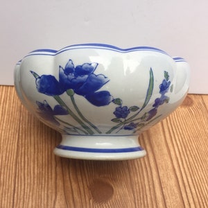 May include: White ceramic bowl with a scalloped rim, decorated with blue floral designs. The bowl has a pedestal base and blue trim. The flowers are painted in shades of blue with green stems and leaves. A decorative piece for the home.