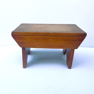 May include: A small wooden stool with a simple design. The stool is made of light brown wood and has a rectangular top and four legs.