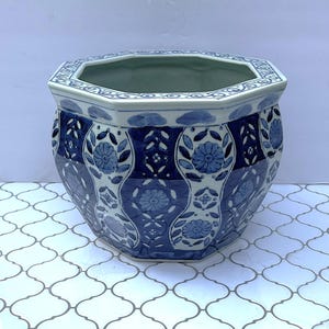 May include: An octagonal ceramic planter with a blue and white floral design. The planter features a repeating pattern of flowers and leaves, with a blue rim and interior. The exterior is decorated with alternating bands of blue and white.