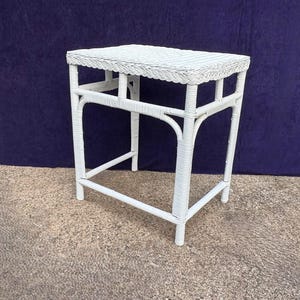 May include: A white wicker side table with a rectangular top and four legs. The table has a decorative woven edge and is painted white. The table is approximately 24 inches tall and 18 inches wide.