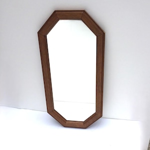May include: A wooden framed mirror with an octagonal shape. The frame is a medium brown color, and the mirror's surface is clear. The mirror is set against a white background.