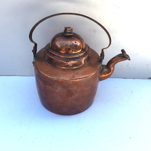 May include: A vintage copper kettle with a rounded body, a hinged lid, and a curved handle. The kettle has a spout on the side and shows signs of age and use.