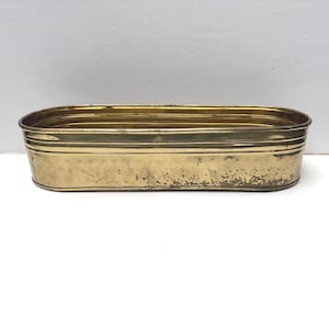May include: An elongated oval-shaped brass planter with a polished metallic finish. The container features horizontal ribbed detailing along the upper rim and shows signs of natural patina and minor surface wear along the lower base section.