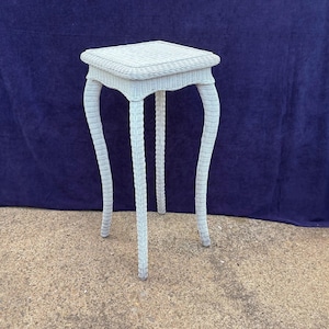 May include: A light blue wicker side table with a square top and curved legs. The table is approximately 28 inches tall and has a woven design on the tabletop and legs. The table is made of wicker.