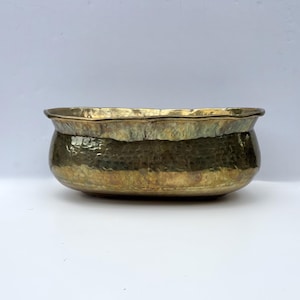 May include: An oval-shaped brass bowl featuring a hammered texture across its exterior surface. The bowl has a wide, smooth rim with a slightly scalloped edge and a deep, rounded basin. The metal displays a warm, aged golden-brown patina.