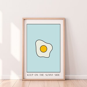 May include: A framed print with a light blue background and a simple line drawing of a fried egg with a yellow yolk. The text below the egg reads 
