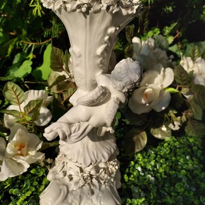 May include: White ceramic candle holder with a cherub figurine and floral details. The holder has a fluted design and a base with a scalloped edge.