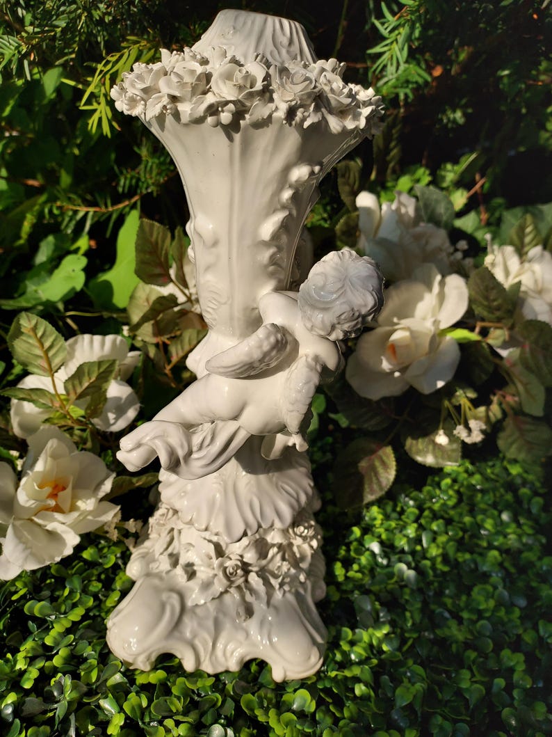 May include: White ceramic candle holder with a cherub figurine and floral details. The holder has a fluted design and a base with a scalloped edge.